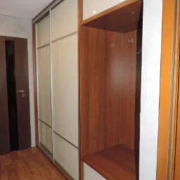 Funkcjonalna szafa wnękowa z przesuwnymi drzwiami i drewnianą wnęką z siedziskiem, oświetlona punktowo, w jasnym wnętrzu z drewnianą podłogą i ciemnymi drzwiami.