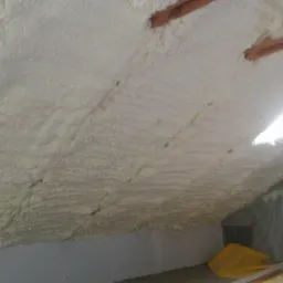 Poddasze po ociepleniu pianką PUR, widoczne drewniane elementy konstrukcji i folia ochronna na ścianie. Biała, gruba warstwa izolacji.