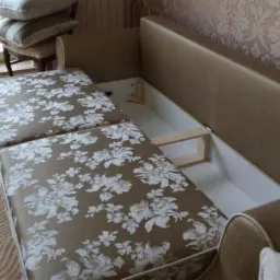 Sofa z otwartym schowkiem, tapicerowana tkaniną w kwiaty. Widoczny mechanizm otwierania i pojemnik na pościel. W tle okno i wzorzysta tapeta.