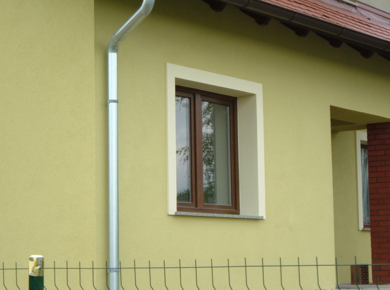 Elewacja budynku z oknem w brązowej ramie, widoczna rynna spustowa i fragment ogrodzenia z siatki.