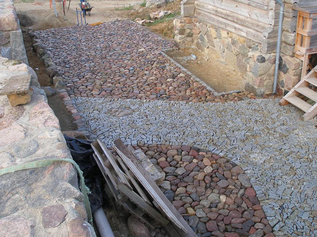 Ułożona ścieżka z kamienia polnego i drobnej kostki granitowej prowadząca do drewnianego domu, z widocznymi elementami konstrukcyjnymi i przygotowanym podłożem.