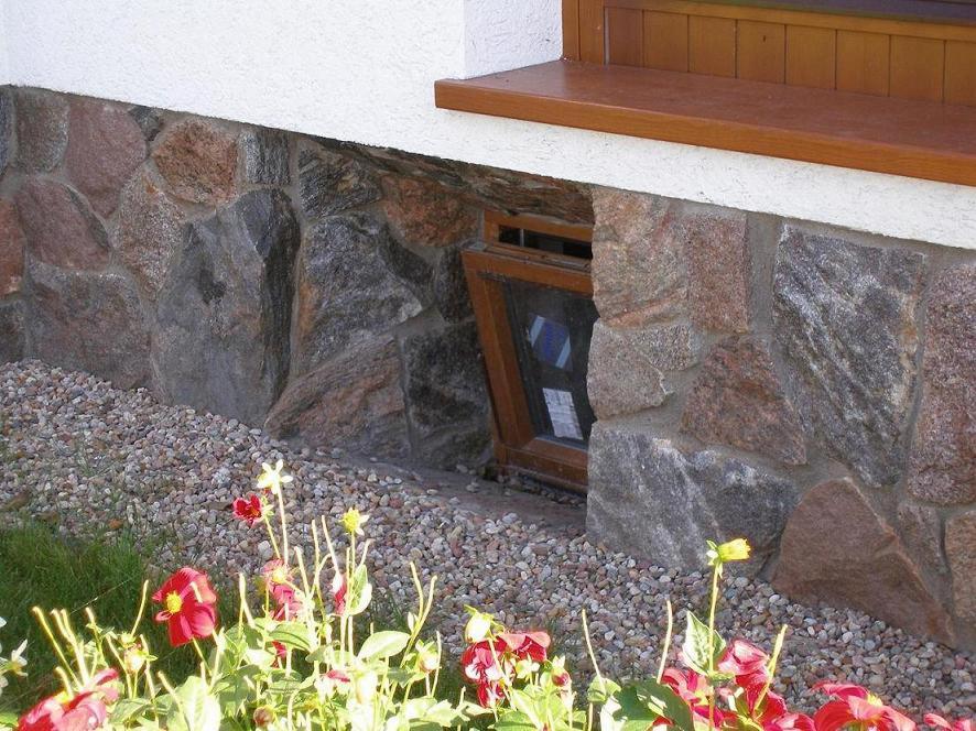 Kamienna elewacja budynku z oknem piwnicznym, ozdobiona drobnym żwirem i kwitnącymi czerwonymi kwiatami na pierwszym planie.