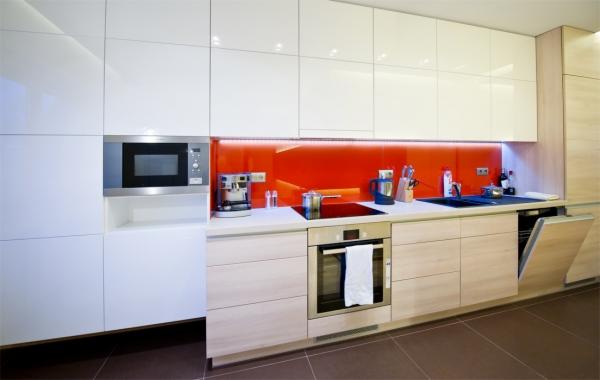 Nowoczesna kuchnia z białymi i drewnopodobnymi szafkami, czerwonym panelem szklanym nad blatem, wbudowaną mikrofalówką i ekspresem do kawy.