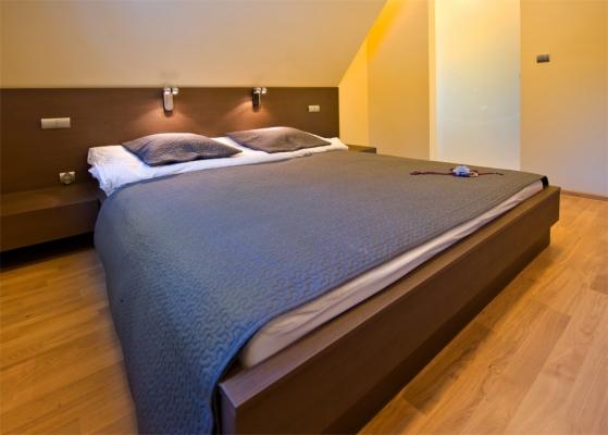 Sypialnia w minimalistycznym stylu z łóżkiem na drewnianej ramie, szarą narzutą i poduszkami; na ścianie lampki nocne.