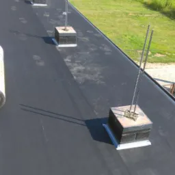 Czarny, płaski dach pokryty papą z trzema kominkami wentylacyjnymi z wystającymi prętami zbrojeniowymi, widoczne ślady stóp na papie.