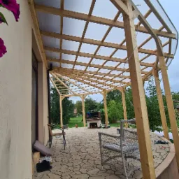 Konstrukcja drewnianej altany z przezroczystym zadaszeniem poliwęglanowym przylegająca do budynku, widok z boku na kamienny taras z meblami ogrodowymi i grillem w tle.