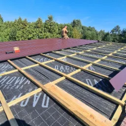 Montaż brązowego pokrycia dachowego na drewnianej konstrukcji więźby, widoczne narzędzia i materiały, w tle zielone drzewa i błękitne niebo.