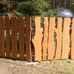 Drewniane ogrodzenie z surowych desek o nieregularnych krawędziach, zamontowane na metalowych słupkach, na tle zieleni i drzew. Widoczny fragment samochodu w tle.