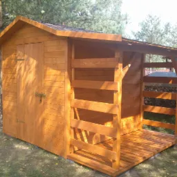 Drewniana altana z zamykanymi drzwiami i zadaszonym miejscem na drewno, wykonana z desek w odcieniach brązu, na tle zielonego trawnika i drzew.