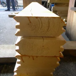 Stos trzech drewnianych belek z wyfrezowanymi krawędziami, przygotowanych do montażu, z widocznymi słojami drewna i śladami kleju, na tle warsztatu.