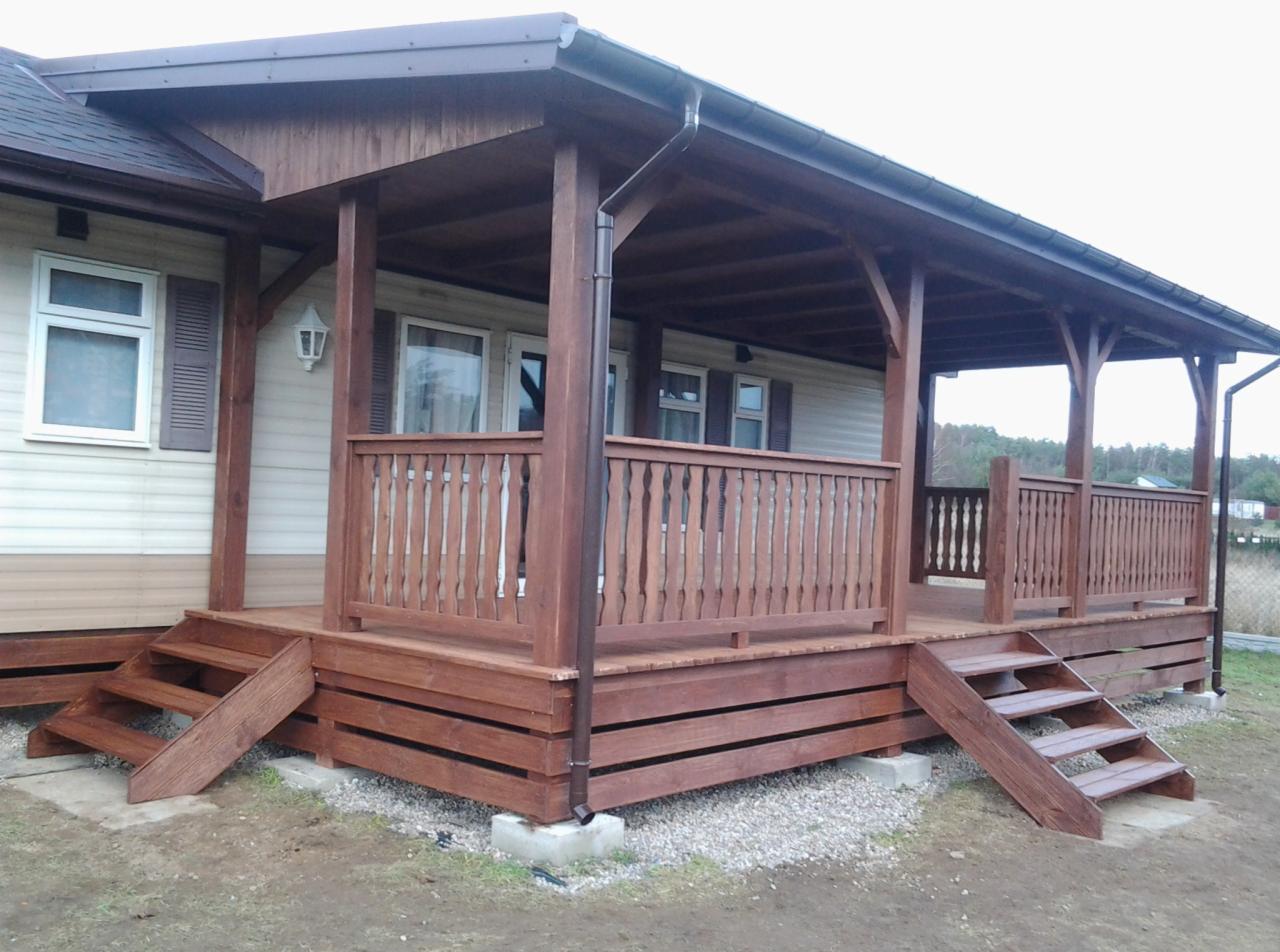 Zadaszony, drewniany taras z balustradą i schodami przy domu z sidingu. Ciemne drewno, jasny siding. Widok z zewnątrz, lekko z dołu.
