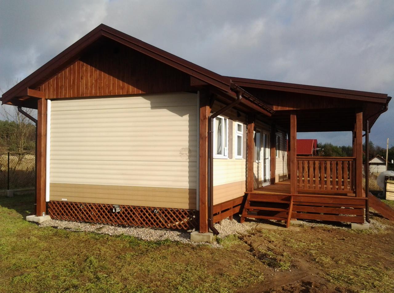 Gotowy dom szkieletowy z drewnianą werandą i beżową elewacją sidingową. Widoczne brązowe elementy wykończeniowe i podmurówka z kratką. Tło: pochmurne niebo.