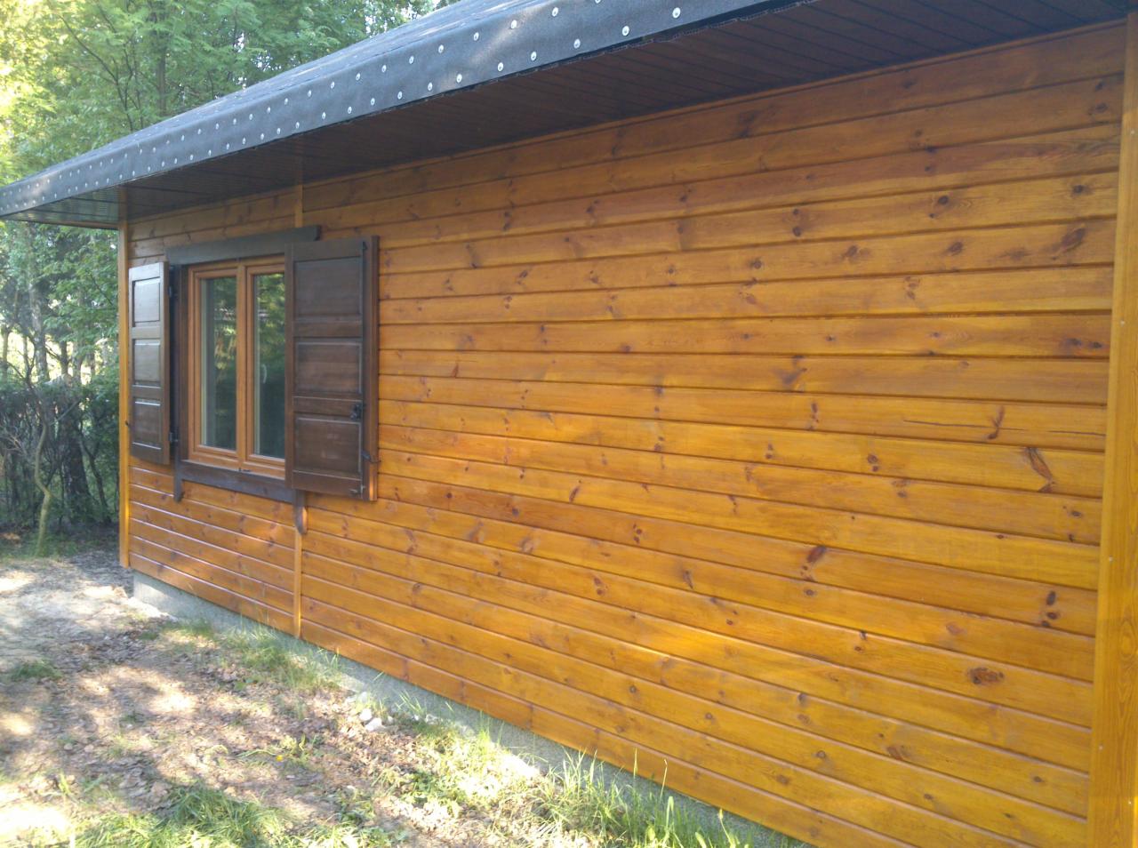 Drewniana altana z oknem i brązowymi okiennicami, widok z boku, oświetlona naturalnym światłem, na tle zieleni. Ściany z wyraźnym rysunkiem słojów.