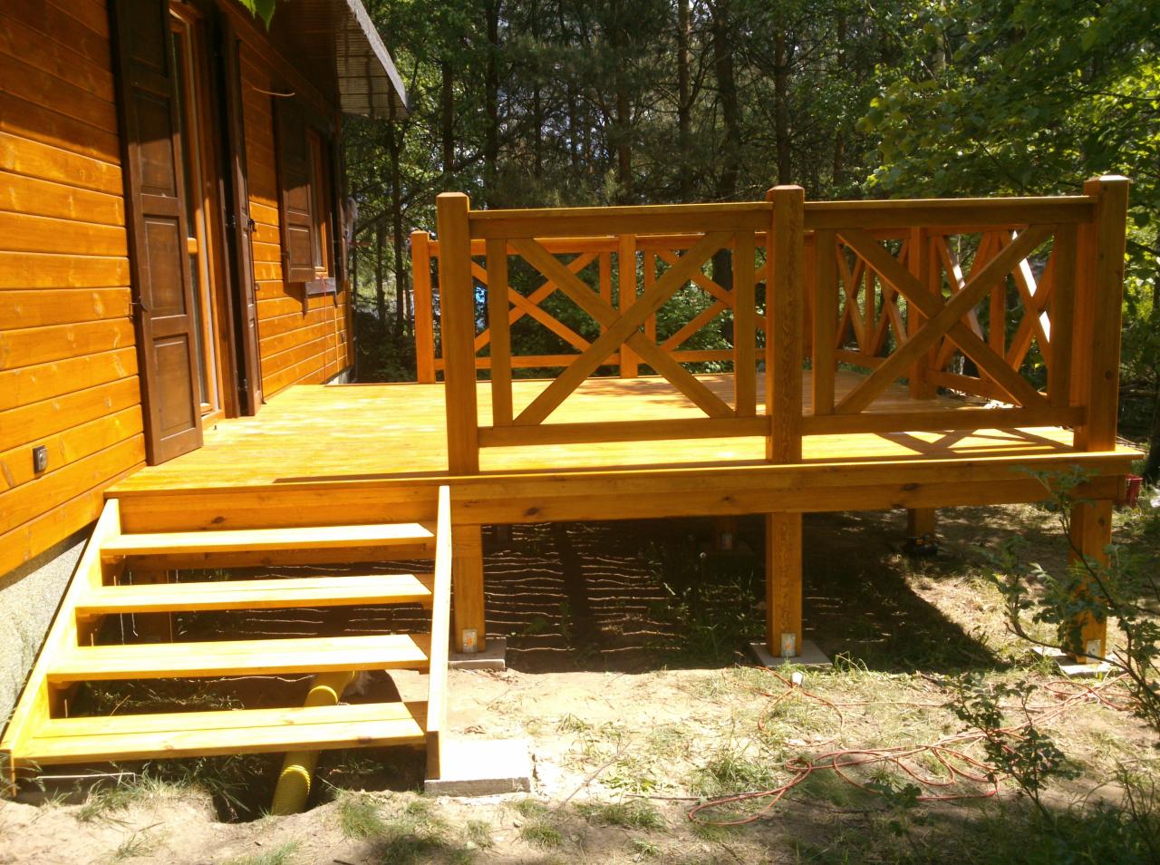 Drewniany taras z balustradą o wzorze krzyżowym, schody wejściowe, deski w ciepłym odcieniu brązu, fragment drewnianego domu, zieleń w tle, słoneczny dzień.