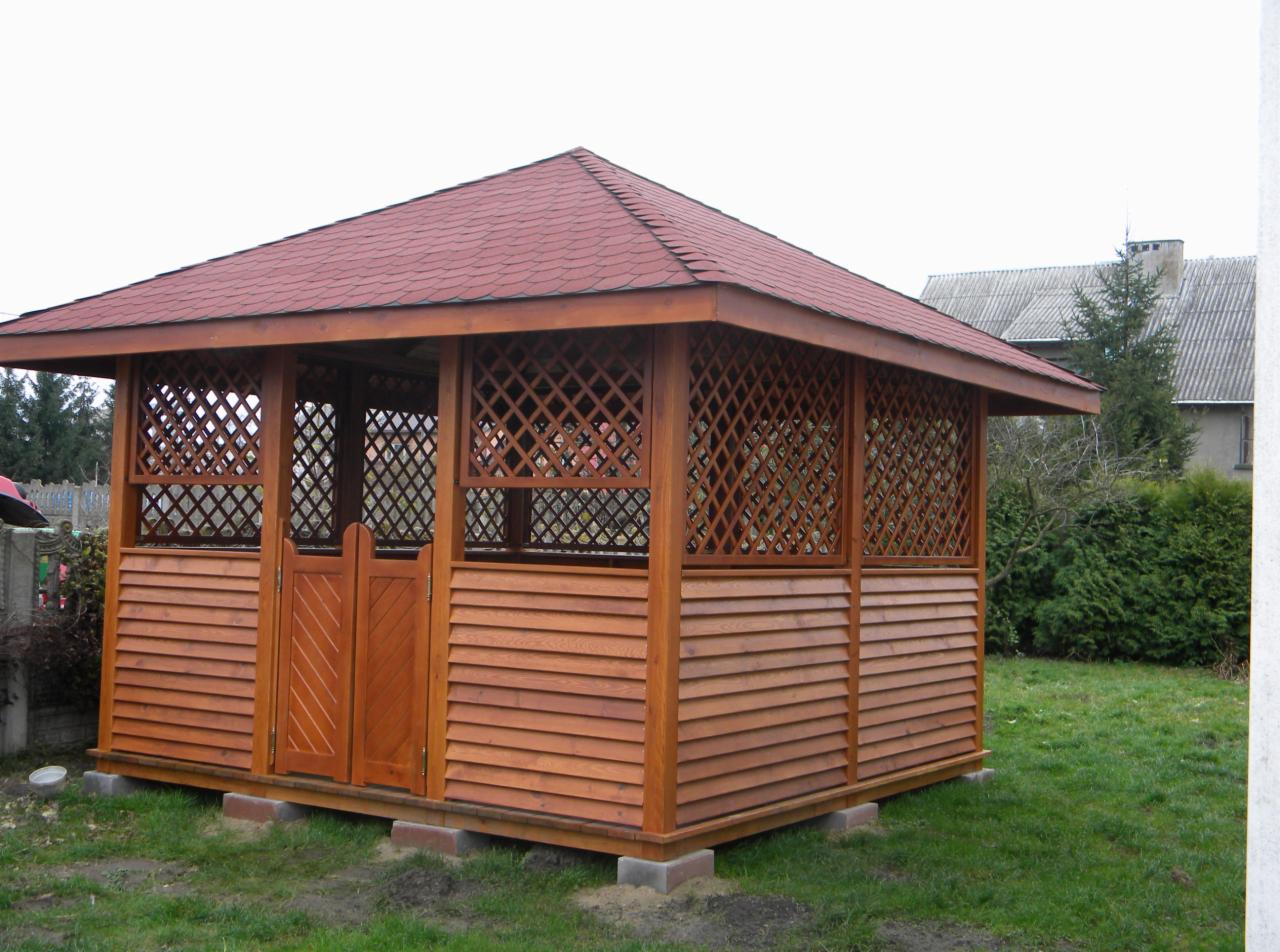 Drewniana altana z kratownicą i dwuskrzydłowymi drzwiami w Nowym Mieście, z czerwoną dachówką, na trawniku. Widok z przodu, w tle budynek mieszkalny.