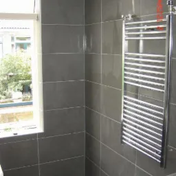 Wykończona łazienka z szarymi, matowymi płytkami wielkoformatowymi na ścianach i podłodze, widoczny grzejnik drabinkowy oraz fragment okna z widokiem na zieleń.