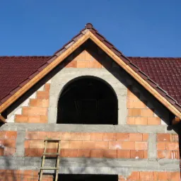 Fragment domu w budowie: czerwona dachówka, ceglana ściana z otworem okiennym w kształcie łuku, drewniana drabina. Błękitne niebo w tle.