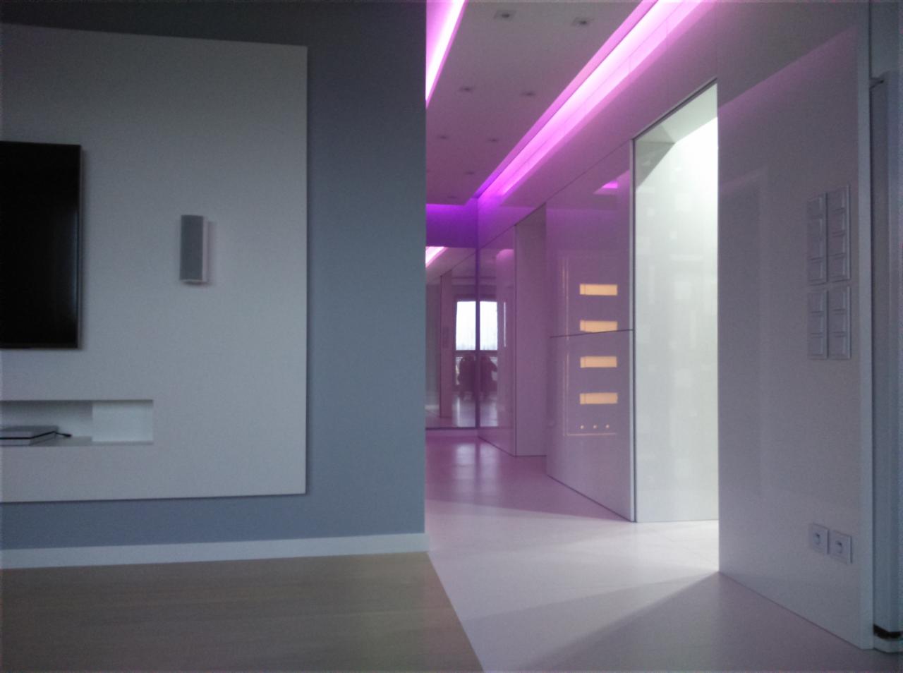 Nowoczesne wykończenie wnętrza: minimalistyczny salon z podświetleniem LED w kolorze fioletowym i zabudową meblową w kolorze białym. Widoczny fragment ściany z telewizorem.