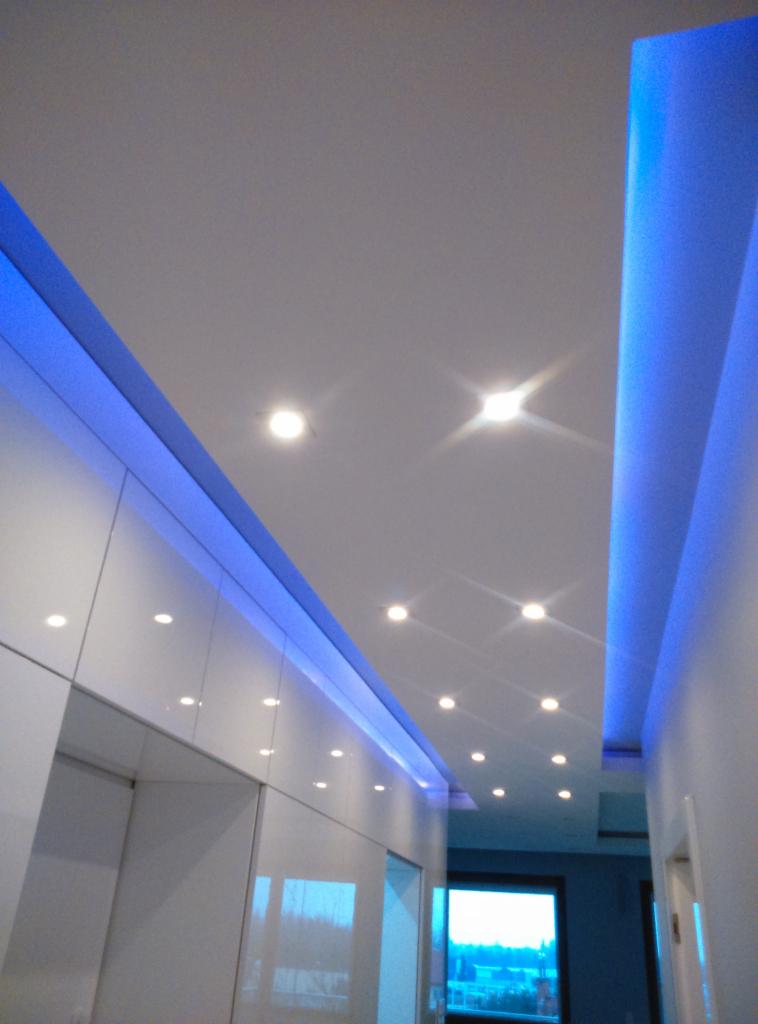 Nowoczesny sufit podwieszany z oświetleniem LED w kolorze niebieskim, punktowe lampy sufitowe, białe ściany i zabudowa meblowa, widok z dołu.