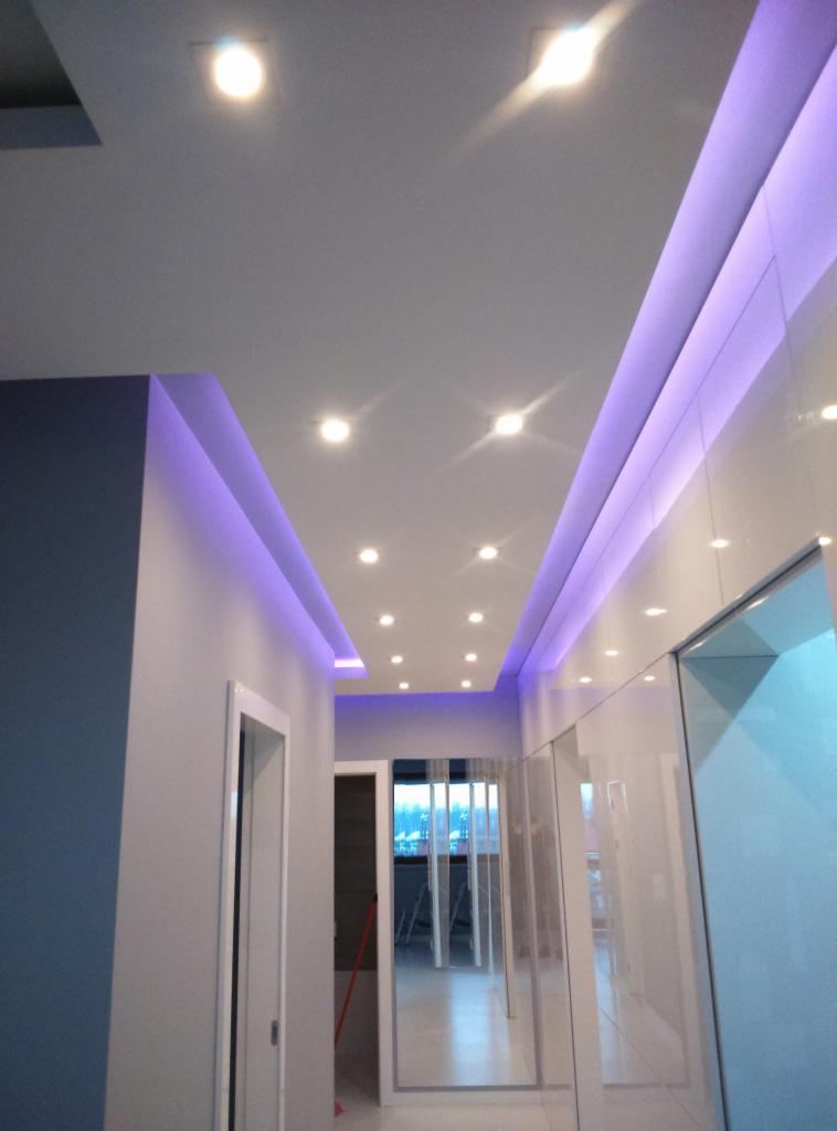 Nowoczesny korytarz z podwieszanym sufitem, oświetleniem LED w kolorze fioletowym i zabudową szaf wnękowych z białymi, błyszczącymi frontami. Widok na wnętrze mieszkania.