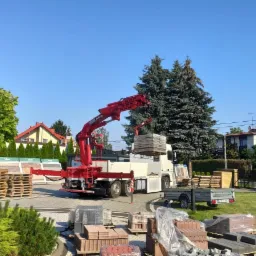 Rozładunek palety materiałów budowlanych za pomocą czerwonego dźwigu samochodowego na placu, widoczne ułożone materiały i przyczepka.