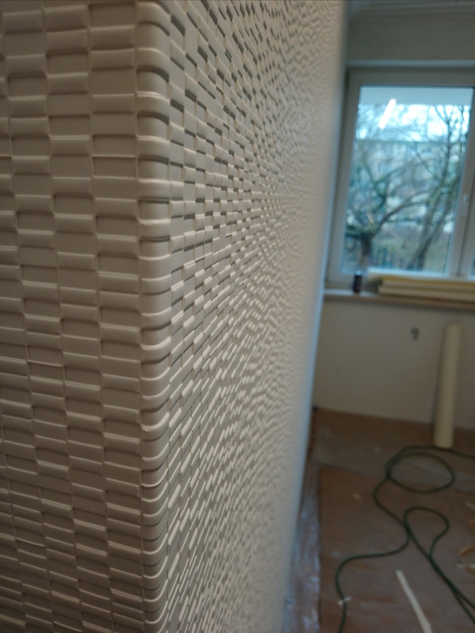 Ściana pokryta trójwymiarową tapetą strukturalną z regularnym wzorem, widok z bliska na róg pomieszczenia, w tle okno i częściowo zakryta podłoga.