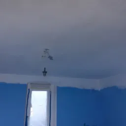 Pokój w trakcie malowania: ściany w kolorze niebieskim częściowo pomalowane białą farbą, okno zabezpieczone folią, widoczne kable oświetleniowe na suficie.