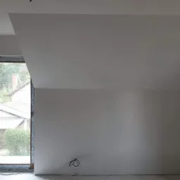 Pokój na poddaszu po remoncie, z jedną ścianą z cegły, świeżo pomalowane ściany na biało, okno dachowe i przewody elektryczne wystające ze ściany.