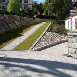 Ułożona ścieżka z kostki brukowej w jodełkę, oddzielona pasem trawy i kamieniami, prowadząca do budynku z kamiennym murem oporowym w tle.