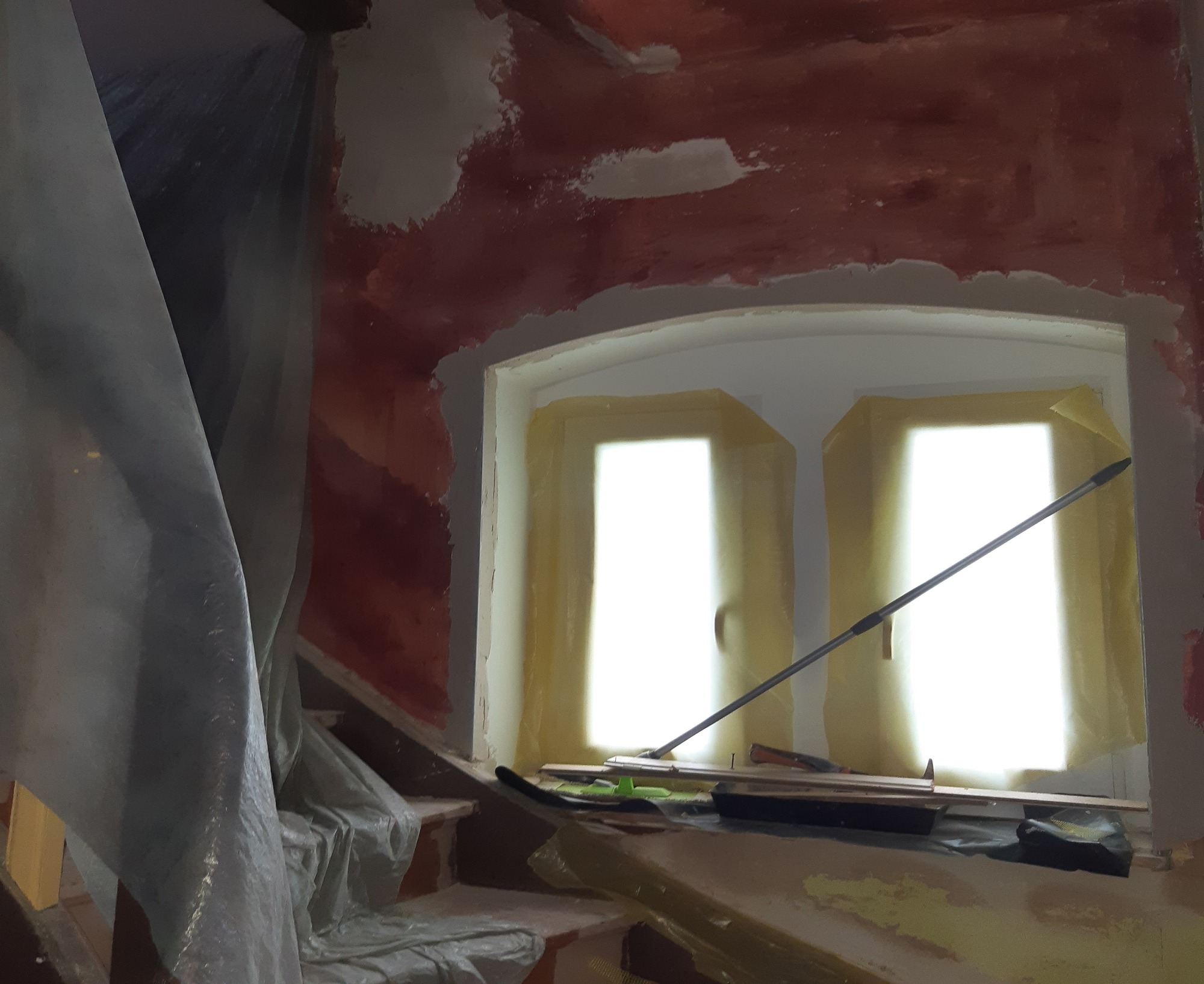 Przygotowania do malowania ścian: ściana z surową farbą, częściowo pokryta gładzią, okno zabezpieczone taśmą malarską, folia ochronna na schodach, narzędzia malarskie w pobliżu.