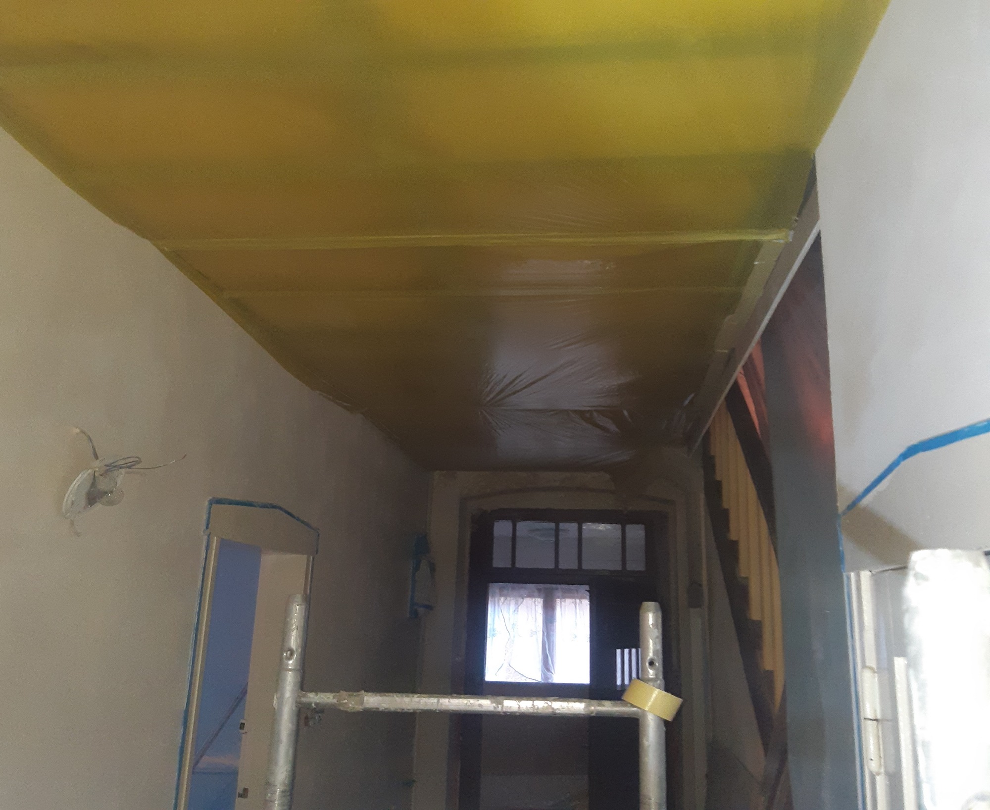 Przygotowanie sufitu do prac wykończeniowych: sufit zabezpieczony folią malarską, widoczne ślady po instalacji elektrycznej, drabina malarska w pomieszczeniu z widocznymi schodami.