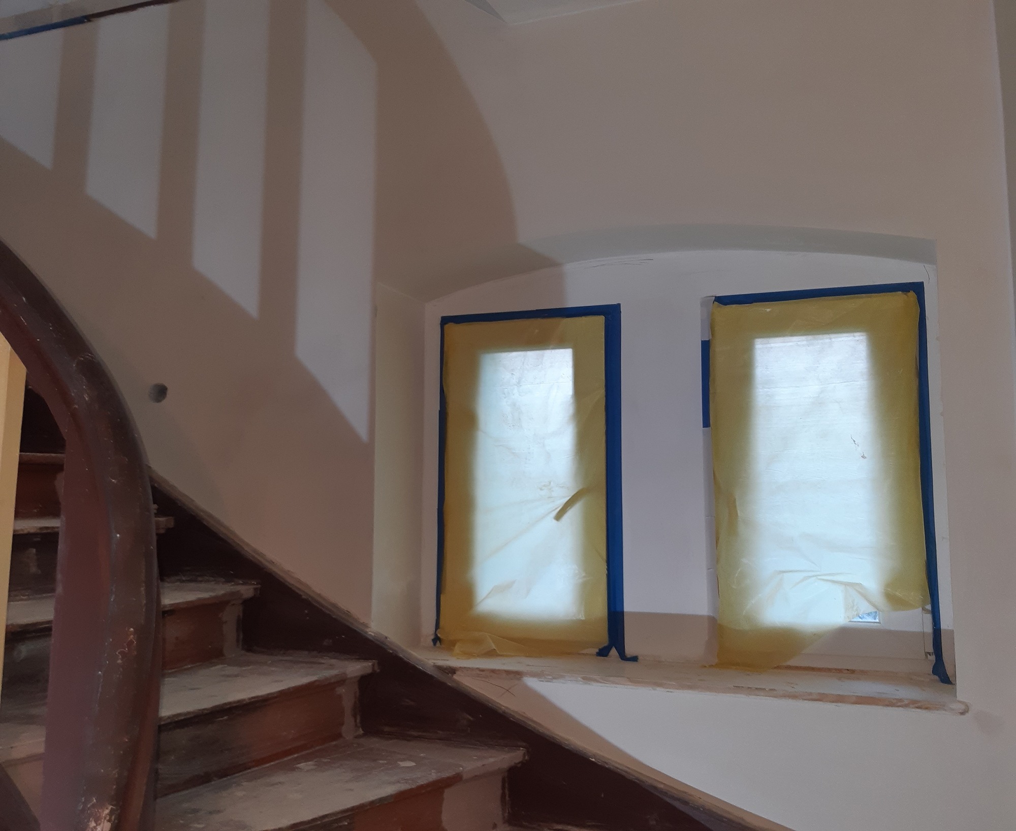 Przygotowanie do malowania klatki schodowej: okna zabezpieczone folią i taśmą malarską, widoczne drewniane schody przed renowacją.