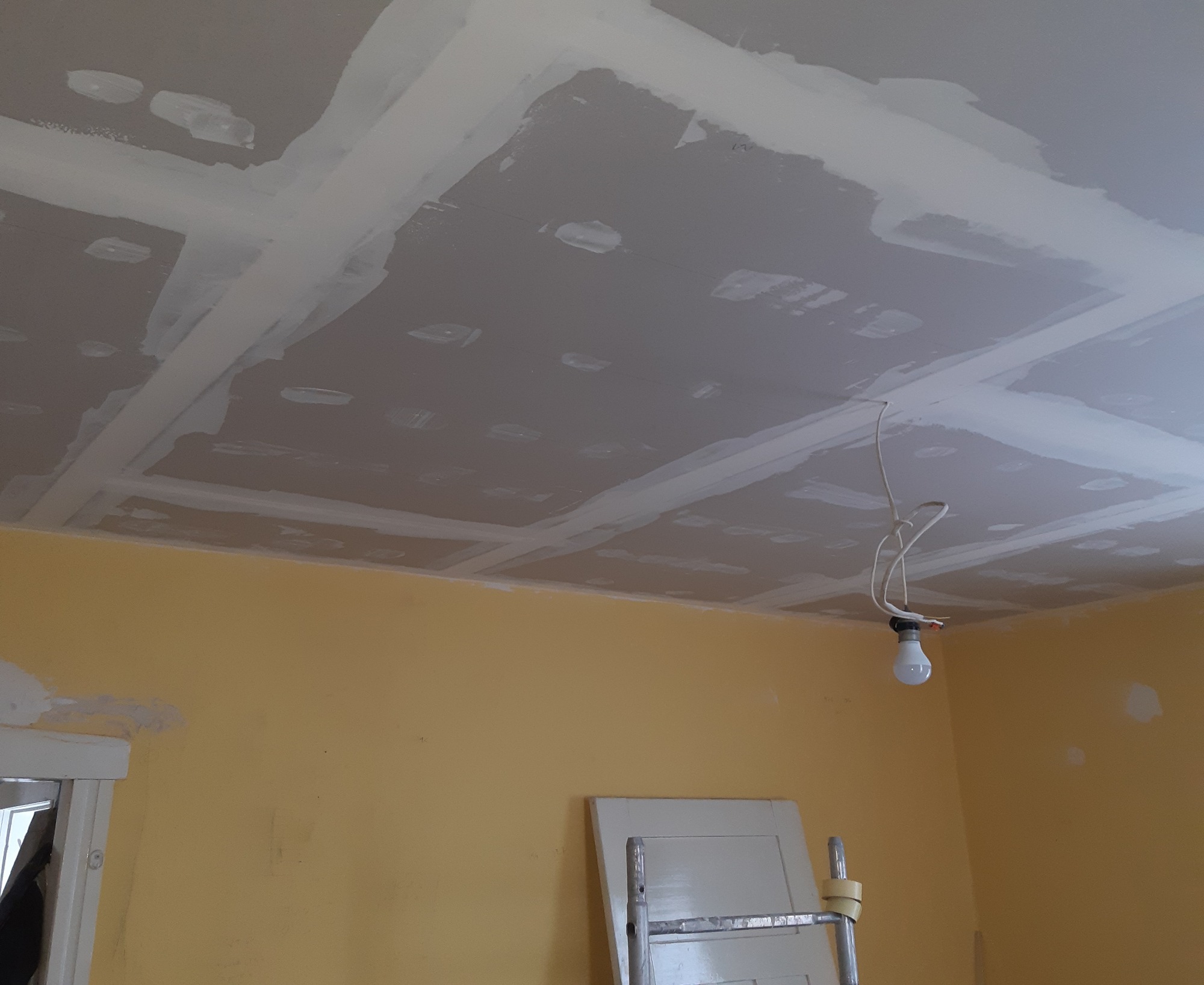 Sufit z płyt kartonowo-gipsowych, z widocznymi łączeniami wzmocnionymi taśmą i pokrytymi szpachlą, w tle żółta ściana z widocznymi śladami po naprawach, wisząca żarówka na kablu.