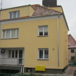 Żółta fasada dwupiętrowego domu z numerem '10', widoczna z boku. Część budynku w trakcie ocieplania, z rusztowaniem. Dach z czerwonej dachówki.