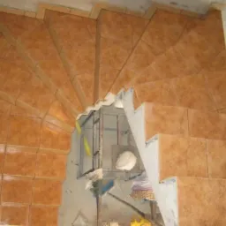 Wykończone schody spiralne z brązowych, kwadratowych płytek ceramicznych. Widok z góry na ułożone płytki, fragment rusztowania w tle.