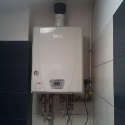 Biały piec gazowy marki Ulrich zamontowany na ścianie wyłożonej biało-czarną glazurą, widoczne liczne rury i zawory podłączone do urządzenia.