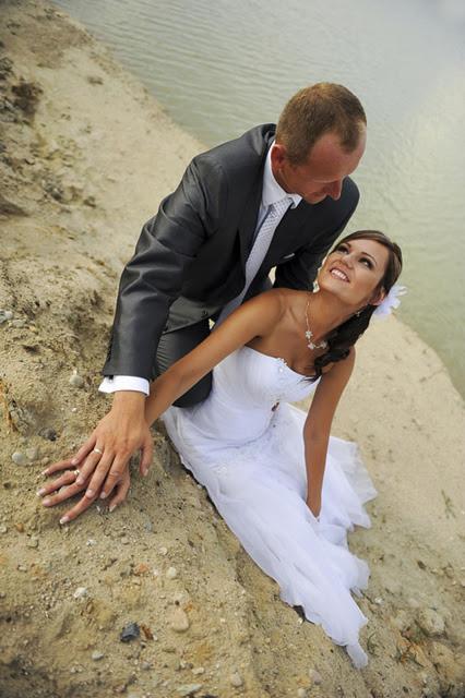 Para młoda w sesji plenerowej: panna młoda w białej sukni spogląda z uśmiechem na pana młodego w szarym garniturze, oboje siedzą na piaszczystym brzegu jeziora, dłonie dotykają piasku.