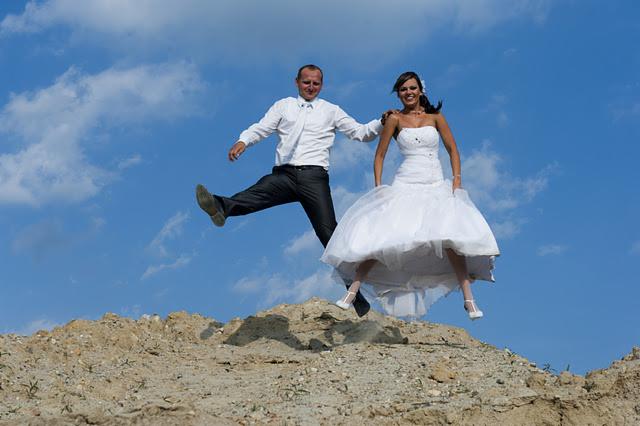 Uśmiechnięta para młoda w białych strojach, skacząca radośnie na szczycie piaszczystego wzgórza na tle błękitnego nieba z chmurami.