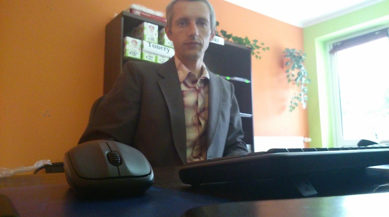 Mężczyzna w marynarce siedzi przy biurku z komputerem w biurze w Dąbrowie Górniczej. Na półce widoczne pudełka z tonerami.