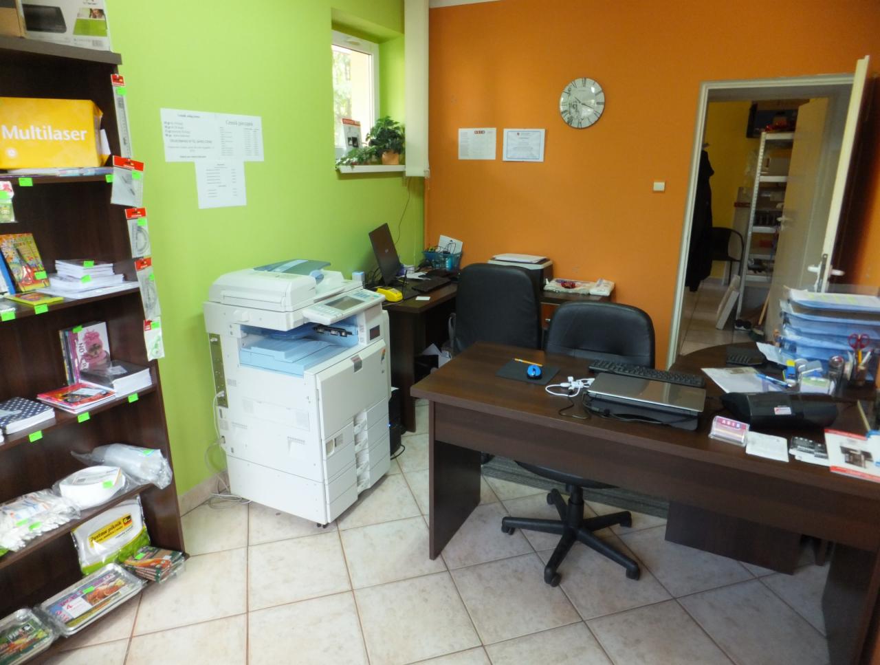 Biuro z pomarańczowymi ścianami, wyposażone w biurka, krzesła, regał z materiałami biurowymi i drukarkę. Widoczne dokumenty i sprzęt komputerowy.