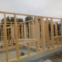 BH MONTER - Surowa drewniana konstrukcja szkieletowa domu na betonowym fundamencie, widoczna piła łańcuchowa i drabina w tle.