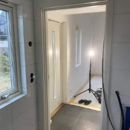 Wnętrze w trakcie remontu: białe płytki na ścianach, nowe szare płytki podłogowe, widoczna lampa budowlana na statywie oraz zabezpieczona folią podłoga w sąsiednim pomieszczeniu.