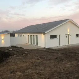 Nowoczesny, parterowy dom z szarym dachem i białymi ścianami, oświetlony zewnętrznymi lampami, widoczny w surowym otoczeniu z nierównym terenem.