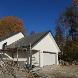 Pracownik na rusztowaniu układa szare dachówki na dachu białego domu z garażem, widok na jesienne drzewa i błękitne niebo.