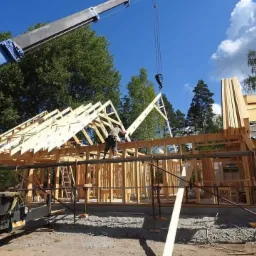 Montaż drewnianej konstrukcji dachu domu jednorodzinnego z użyciem dźwigu, widoczne rusztowania i elementy konstrukcyjne ścian.