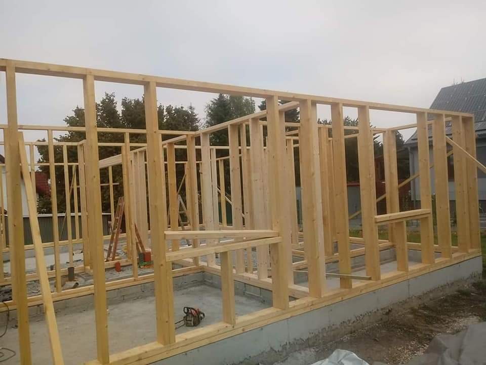 Surowa drewniana konstrukcja szkieletowa domu na betonowym fundamencie, widoczna piła łańcuchowa i drabina w tle.