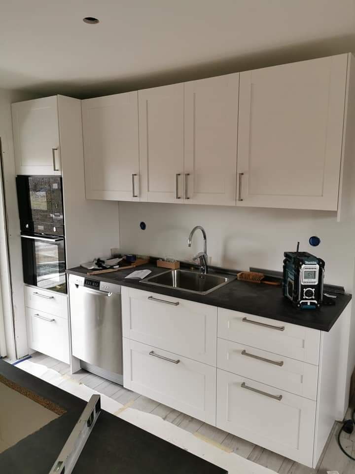 Wnętrze kuchni w trakcie montażu z białymi szafkami, ciemnym blatem, zlewozmywakiem i zainstalowaną zmywarką. Na blacie widoczne narzędzia i radio budowlane.