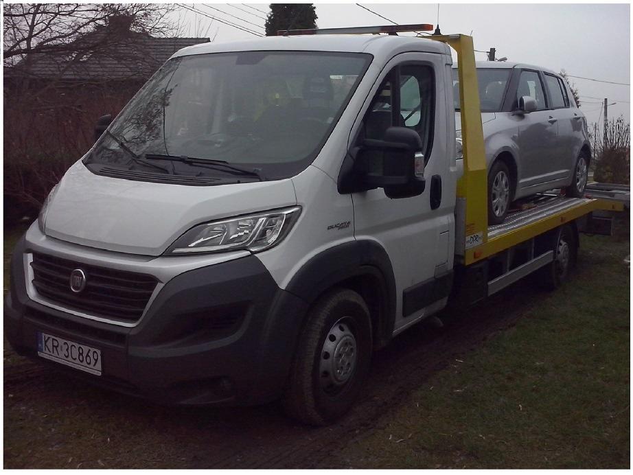 Laweta Fiat Ducato z żółtym najazdem transportuje srebrny samochód osobowy na tle zielonej trawy i drzew.