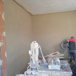 Pracownik wyrównuje świeżo nałożony tynk na ścianie za pomocą szlifierki, widoczna maszyna do tynkowania, schody w tle.