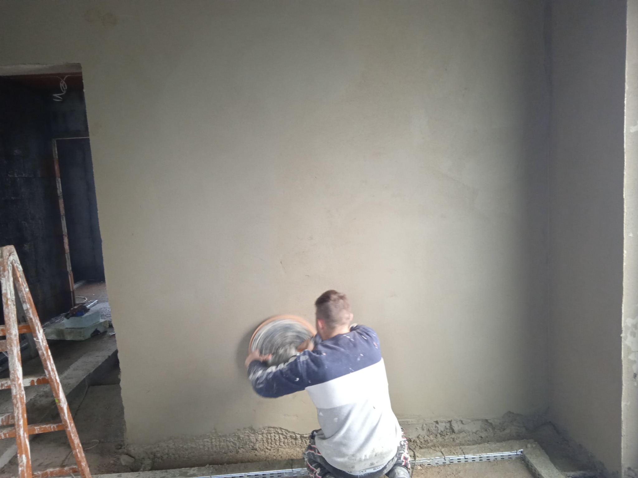Pracownik szlifuje świeżo nałożony tynk na ścianie za pomocą szlifierki rotacyjnej, widoczna drabina i surowy stan wykończenia pomieszczenia.
